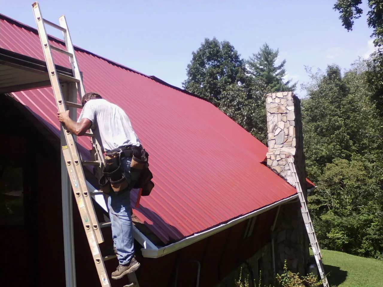 Roofer on a ladder beside a red metal roof and stone chimney — Soffit & Fascia Repair by Roofing Brothers in Singers Glen